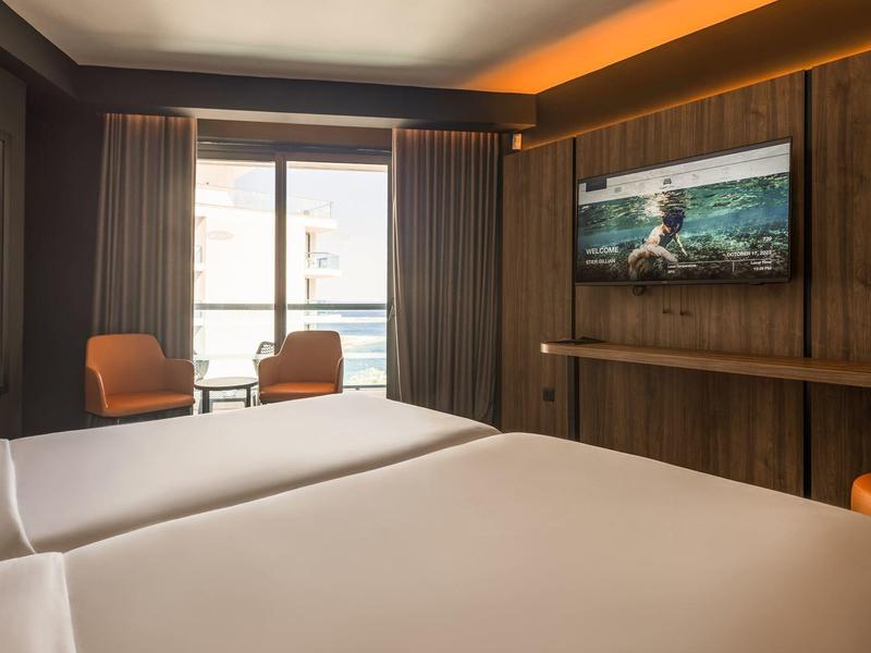 Modernes Hotelzimmer mit zwei Betten, einem Fernseher, Sesseln und großem Fenster mit Vorhängen.