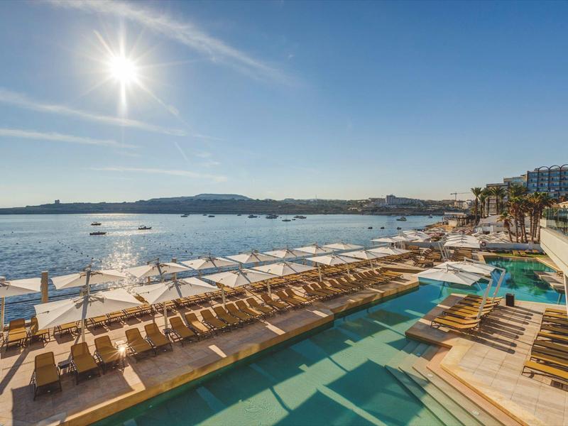 Blick auf eine Strandpromenade mit Liegestühlen, Sonnenschirmen und einem Pool bei sonnigem Wetter.