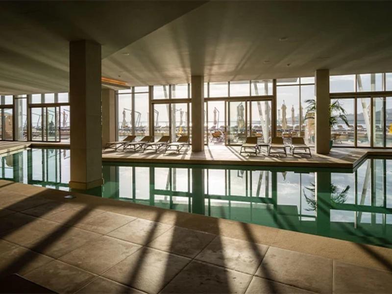 Innenpool mit Liegestühlen und großen Fenstern, durch die Sonnenlicht strömt.