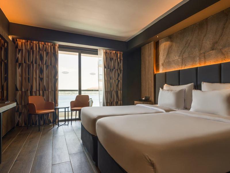 Modernes Hotelzimmer mit zwei Betten, Stuhl, Tisch und Fenster mit Blick nach draußen.