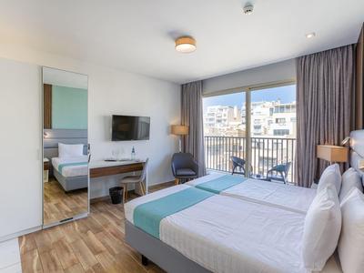 Modern hotelkamer met bed, bureau, tv en balkon met uitzicht op omringende gebouwen.