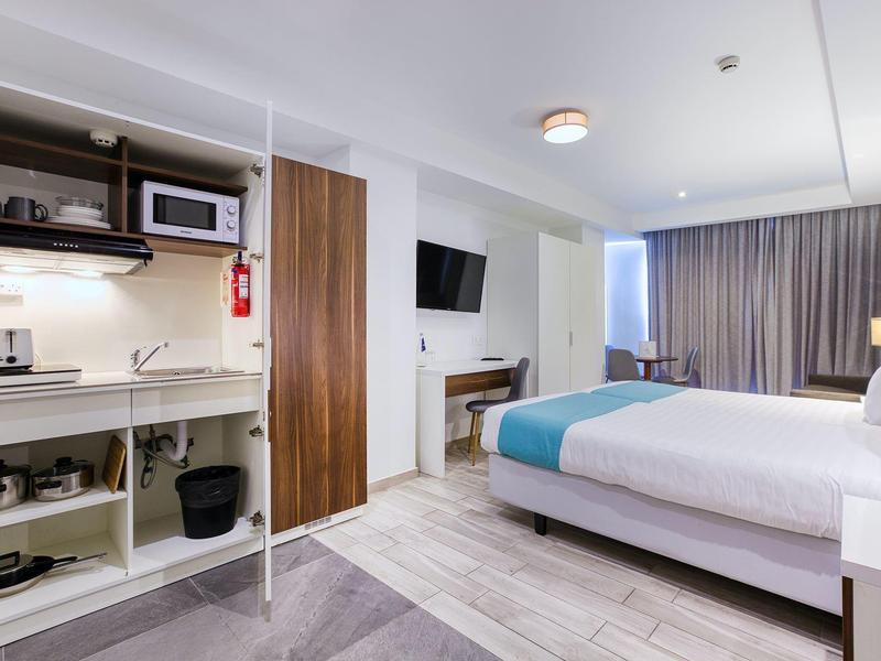 Modern hotelkamer met tweepersoonsbed, wand-tv en open kitchenette in neutrale kleuren.