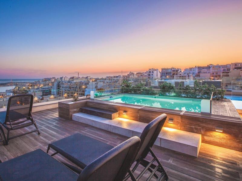Dakterras met zwembad en ligstoelen met uitzicht op stad en zee bij zonsondergang.
