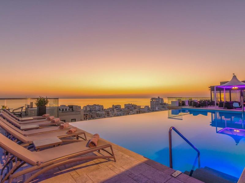 Basen typu infinity z leżakami i widokiem na zachód słońca nad morzem.