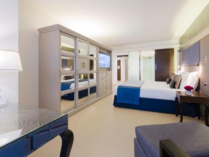 Nowoczesny pokój hotelowy z dużym łóżkiem, niebieskimi akcentami i lustrzanymi szafami.