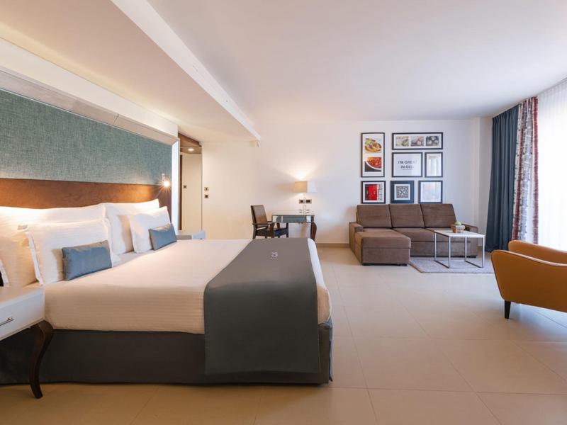 Jasny pokój hotelowy z dużym łóżkiem, sofą, biurkiem i żółtym fotelem.