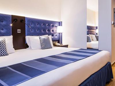 Modernes Hotelzimmer mit Doppelbett, blauer Bettdecke und stilvollem Kopfteil aus Polsterstoff.