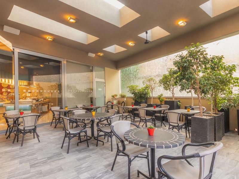 Gemütlicher Außenbereich eines Hotels mit Tischen, Stühlen und Pflanzen unter einem Glasdach.