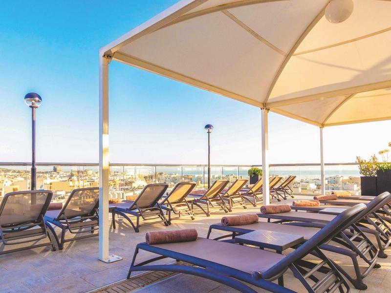 Sonnenliegen unter einem Sonnensegel auf einer Hotelterrasse mit Blick auf die Stadt.