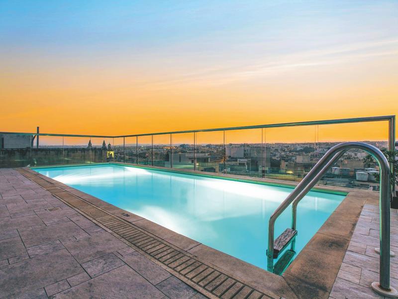 Ein rechteckiger Pool auf einer Dachterrasse mit Blick auf die Stadt bei Sonnenuntergang.