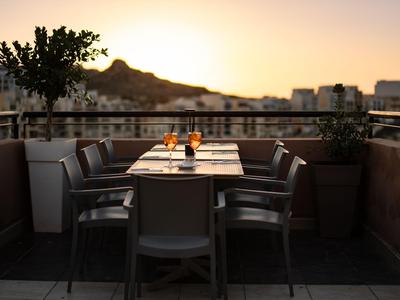 Abendliche Terrasse mit Tisch, sechs Stühlen und Getränken vor Bergkulisse bei Sonnenuntergang.