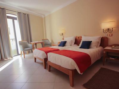 Helles Hotelzimmer mit zwei Einzelbetten, roten Decken und Balkonzugang.