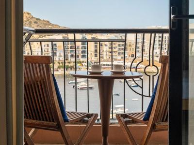 Balkon mit Holztisch und zwei Stühlen mit Blick auf Boote und Gebäude an der Küste.