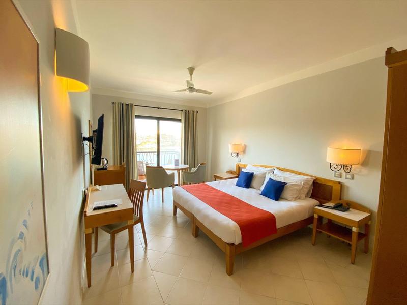 Bright hotel room with a double bed, desk, and balcony overlooking the sea.
