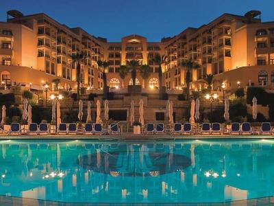 Großes luxuriöses Hotel mit beleuchtetem Pool und Palmen in warmem Abendlicht.