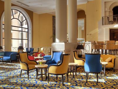 Ruime hotel lounge met gele muren, blauwe en gele stoelen en grote ronde ramen.