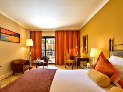 Helles Hotelzimmer mit großem Bett, orangefarbenen Vorhängen und gemütlicher Sitzgruppe.