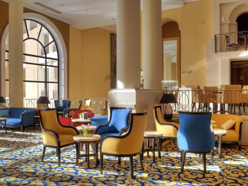 Ruime hotel lounge met gele muren, blauwe en gele stoelen en grote ronde ramen.