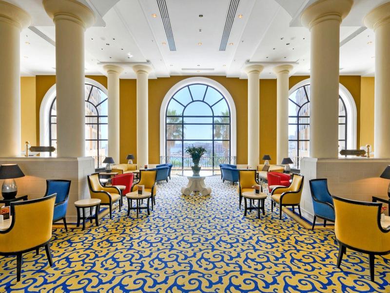 Elegante hotel/lounge met gele muren, grote zuilen en een blauw-gele patroon tapijt.