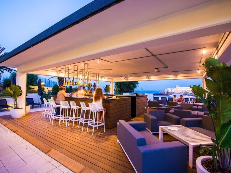 Gezellige hotel dakterraslounge met bar, zithoeken, planten en sfeerverlichting onder een overdekt luifel.