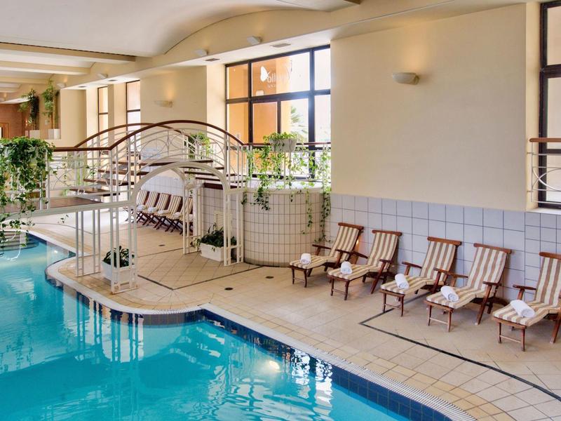 Overdekt hotelzwembad met waterstralen, ligstoelen en planten bij grote ramen.