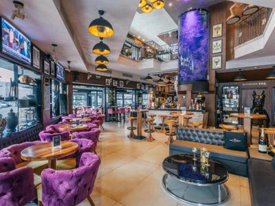 Modern eingerichtete Hotelbar mit violetten Sesseln, Theke und großem Display an der Decke.