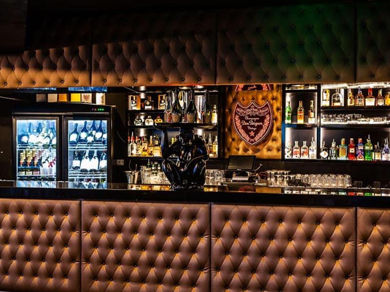 Ciemny bar hotelowy z pikowanym barem i oświetlonymi półkami pełnymi butelek.