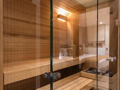 Pokój sauny z drewnianymi ścianami, szklaną frontową ścianą, ławkami i ściennym światłem.