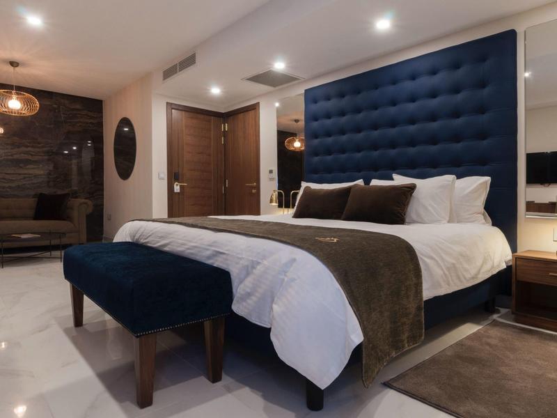 Moderní hotelový pokoj s velkou modrou postelí, lavičkou a teplým osvětlením.