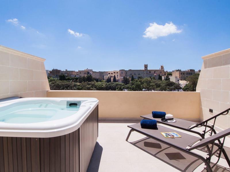 Terasa s vířivkou, dvěma lehátky a výhledem na město pod modrou oblohou.