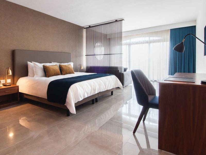 Moderní hotelový pokoj s velkou postelí, psacím stolem, židlí a okny od země ke stropu.