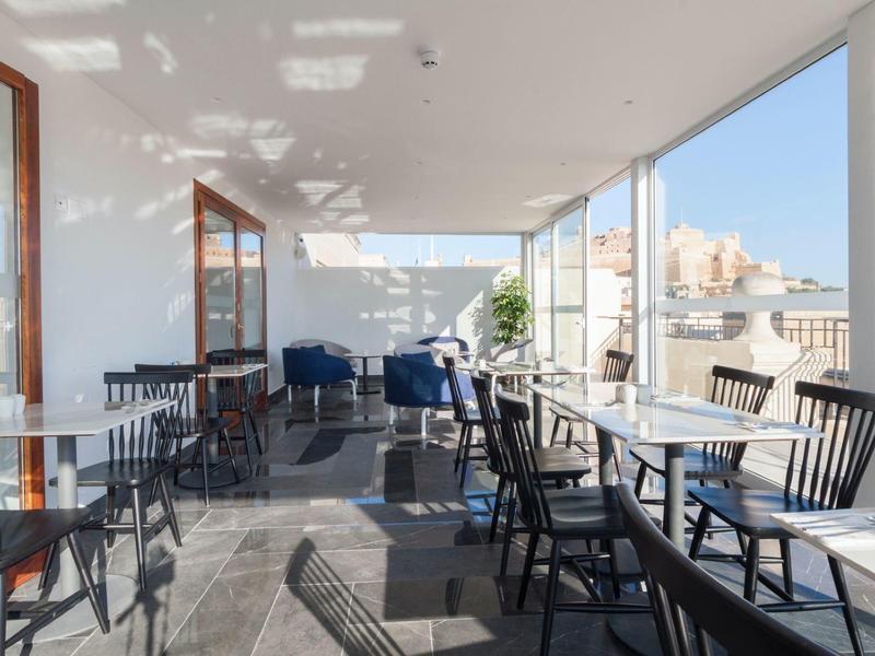 Světlá terasa s židlemi a stoly, výhled na město a moře v pozadí.