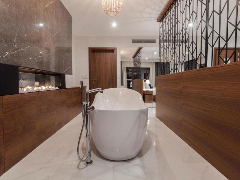 Moderní bílá volně stojící vana mezi dřevěným nábytkem a kamenným obkladem.