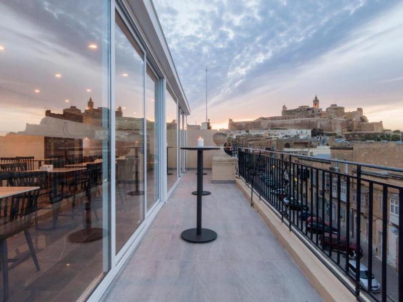 Balkon s výhledem na město a hrad při západu slunce, se stoly a židlemi.