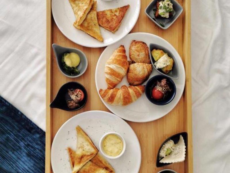 Dřevěný tác s croissanty, trojúhelníkovými sendviči a malými misky s omáčkami a přílohami.