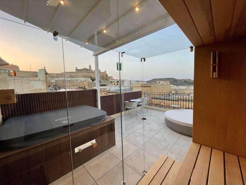 Terasa se skleněnými stěnami, lavičkou a lehátkem s výhledem na město při západu slunce.