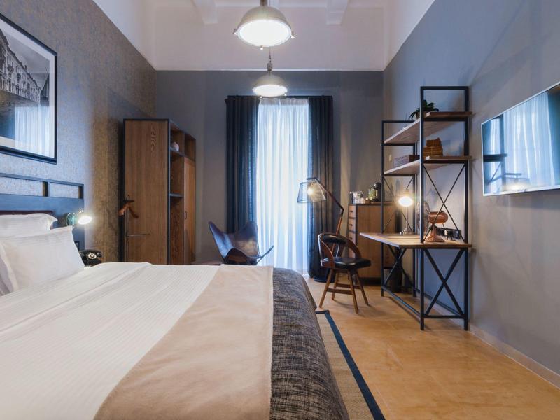 Nowoczesny pokój hotelowy z łóżkiem podwójnym, biurkiem i telewizorem na ścianie w neutralnych kolorach.