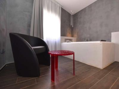 Modernes Hotelzimmer mit grauen Wänden, schwarzem Sessel, rotem Beistelltisch und weißem Sofa.