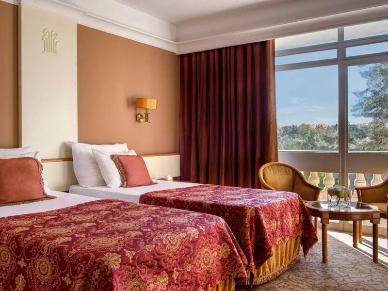 Hotelzimmer mit zwei Einzelbetten, roten Bettdecken, Tisch mit Stühlen und großem Fenster mit Blick ins Grüne.