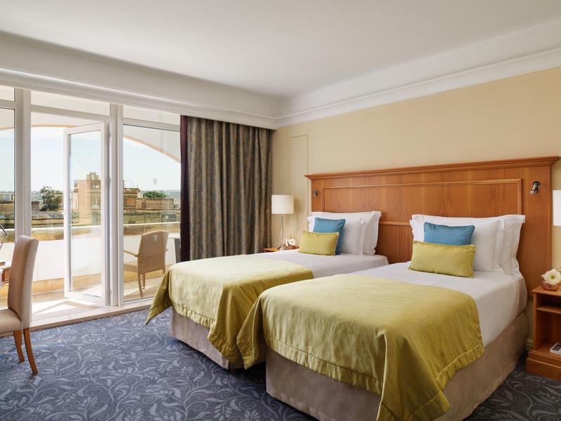 Hotelzimmer mit zwei Einzelbetten, Holzkopfteil, blauen Teppich und Balkon mit Aussicht.