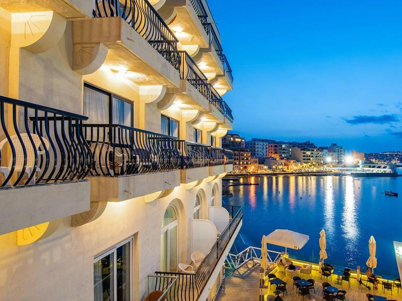 Widok na oświetlony hotel z balkonami nad morzem i spokojny port o zmierzchu.