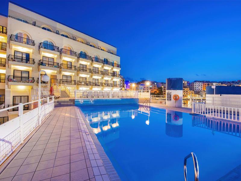 Oświetlony basen hotelowy z widokiem na balkony przy czystym wieczornym niebie.