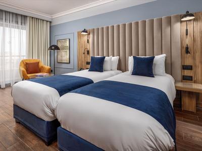 Modernes Hotelzimmer mit zwei Einzelbetten, blauen Akzenten und gelbem Sessel.
