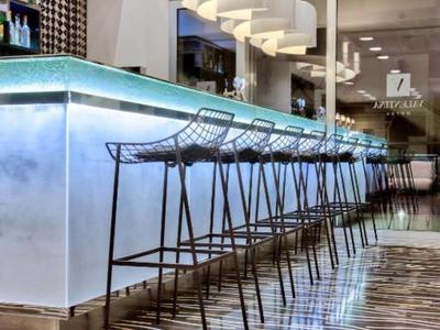 Área de bar moderna en hotel con mostrador alto, taburetes metálicos y iluminación elegante.