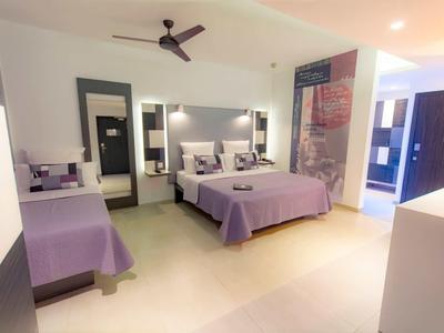 Habitación de hotel luminosa con dos camas, ropa de cama morada, gran espejo y ventilador de techo.