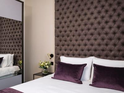Cozy hotel bed with white sheets, purple pillows, tufted headboard, nightstand, and flowers.