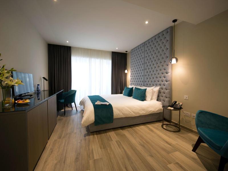 Modern hotel room with a double bed, blue accents, wooden floor, and natural light from a large window.