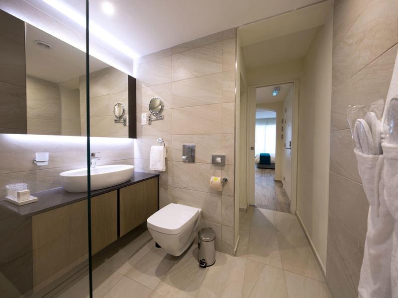 Modern hotel bathroom with beige tiles, sink, toilet, mirror, and bathrobe on the door.