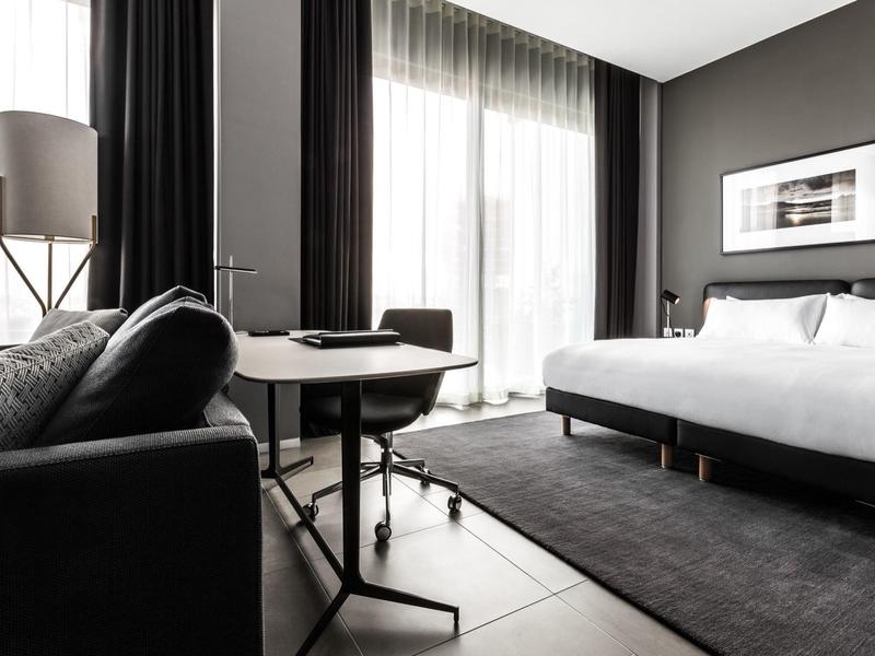 Modern eingerichtetes Hotelzimmer mit großem Bett, Sofa und großem Fenster mit Vorhängen.