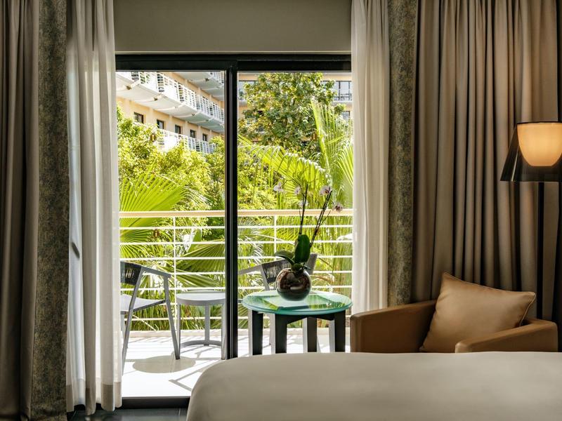 Hotelzimmer mit Balkon, Sesseln und Tisch mit Vase, Blick auf grünes Laub und Gebäude.